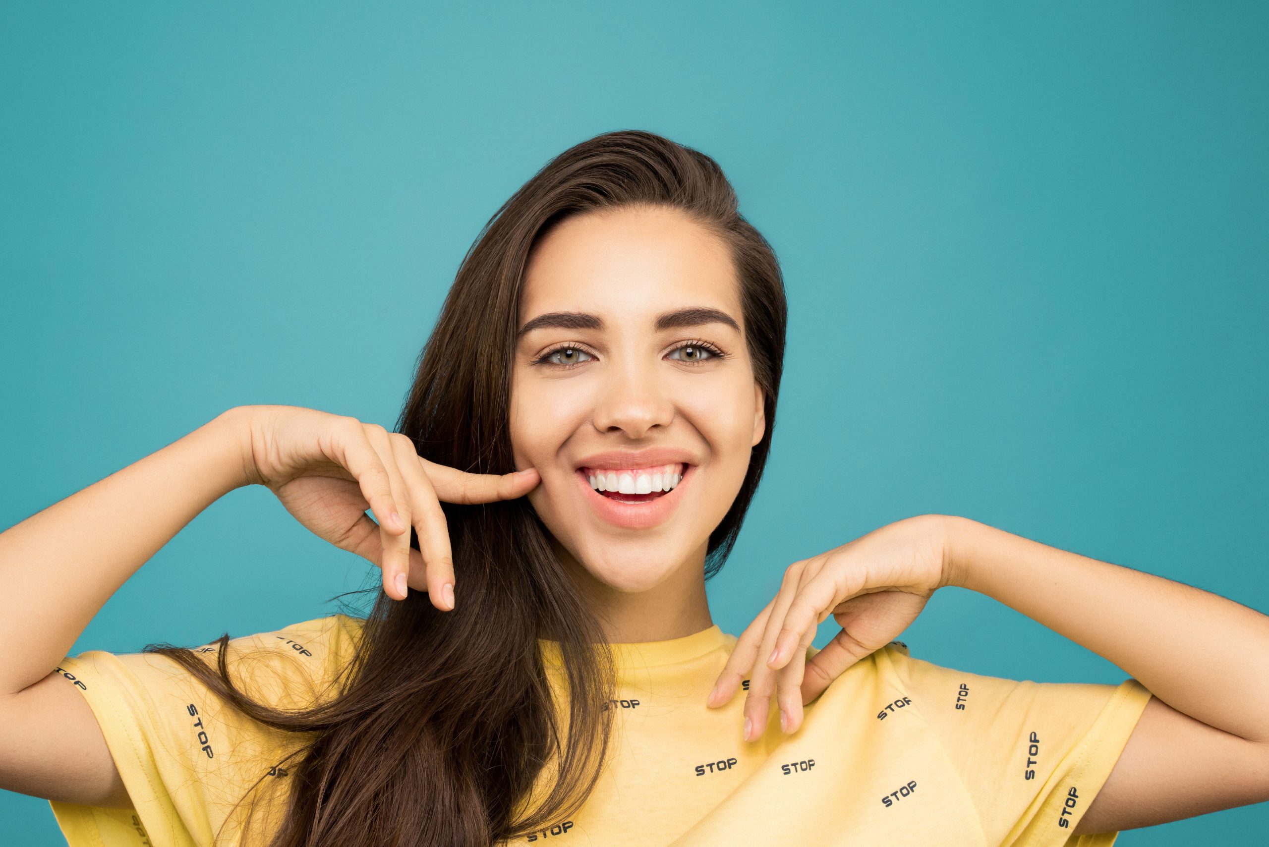 Smiling woman in a yellow shirt pointing to her white teeth against a teal background